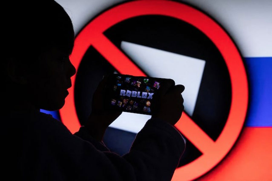 A boy poses for a photo while holding a smartphone in front of a screen displaying a crossed-out logo of the U.S. children's gaming platform Roblox against the background of the Russian state flag, in this illustration taken December 8, 2025.