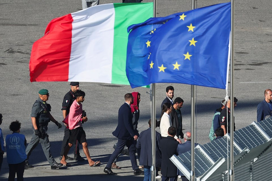 Migrants and security officials walk next to the European Union flag and Italian flag as they disembark from the Italian navy ship Libra that arrived in Albania as part of a deal with Italy to process thousands of asylum-seekers caught near Italian waters, in Shengjin, Albania, October 16, 2024.